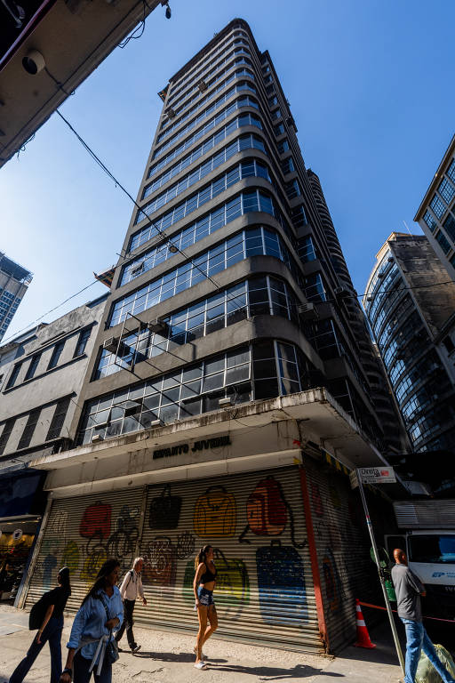 Prédio comercial alto com fachada de vidro e estrutura preta, visto de baixo para cima em dia claro. Na calçada, quatro pedestres caminham próximos à entrada com portas metálicas fechadas e grafites coloridos. Edifícios vizinhos e céu azul completam o cenário urbano.