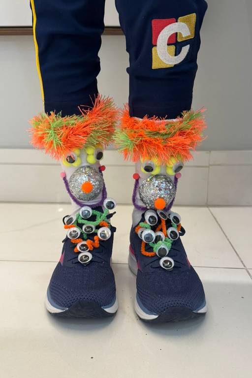 Tênis azul escuro com detalhes em branco, decorados com olhos de brinquedo e botões coloridos. Tornozelos envoltos por pelúcia laranja e verde neon, com bolas prateadas e olhos fixados na parte frontal.