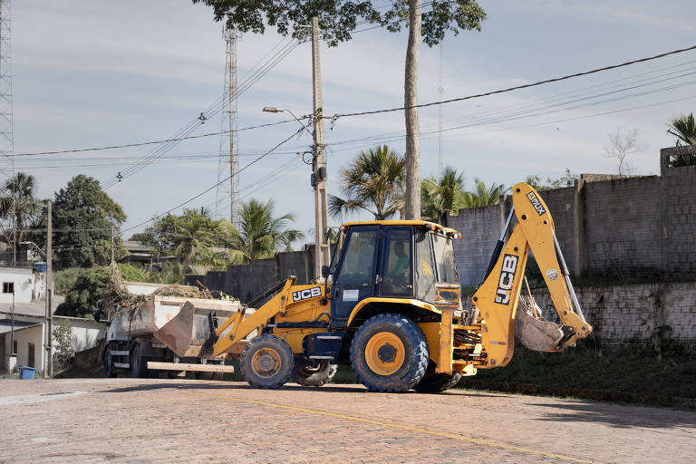 Retroescavadeira amarela da marca JCB estacionada em rua pavimentada, com caçamba dianteira e braço escavador traseiro. Ao fundo, muro de blocos de concreto, árvores e postes com fiação elétrica visíveis sob céu claro.