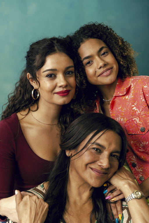 Três mulheres posam juntas sorrindo contra um fundo azul. Duas estão em pé atrás, uma com blusa vermelha e brincos de argola, outra com camisa laranja estampada. A terceira está à frente, apoiando o queixo na mão, usando blusa colorida e relógio prateado.