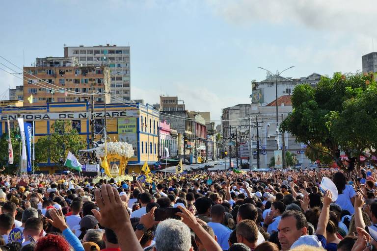 Multidão acompanha procissão religiosa em rua de cidade, com pessoas de costas e algumas erguendo as mãos. Ao fundo, edifícios comerciais e residenciais, incluindo um prédio com letreiro 'Senhora de Nazaré'. Elementos da procissão incluem estandartes e uma estrutura dourada decorativa.