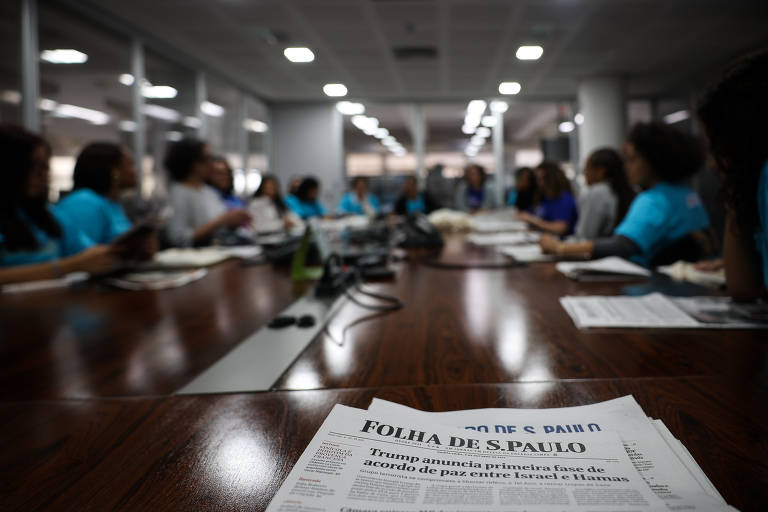 Mesa de conferências com várias pessoas sentadas ao redor, algumas vestindo camisetas azuis. Em primeiro plano, jornal Folha de S.Paulo com manchete sobre Trump anunciando acordo de paz entre Israel e Hamas. Ambiente interno com iluminação artificial e foco no jornal.