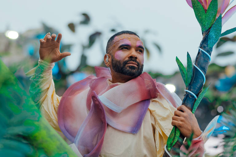 Homem com barba e cabelo curto usa fantasia com tecido translúcido rosa e bege, segurando adereço verde e rosa. Fundo desfocado sugere ambiente externo com outras pessoas.
